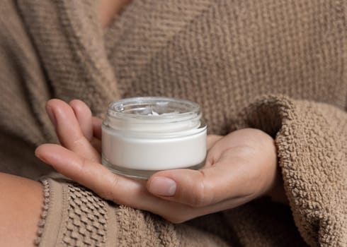 Close up of woman in light brown bathrobe holding white opened cream jar in hands with natural manicure, cosmetic mockup. Bodycare concept. Cropped Image
