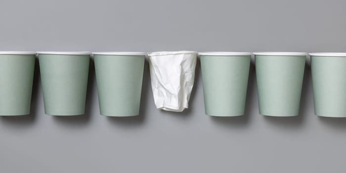 Disposable new light green paper cups and crumpled used white one in a line a gray top view, mockup with copy space. Empty cardboard coffee cups flat lay. No plastic and sustainable lifestyle concept