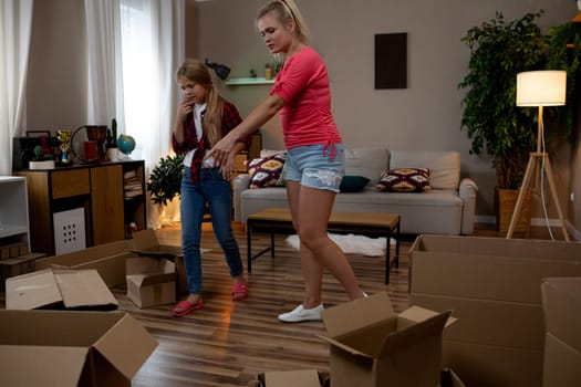 Two cheerful girls are joyfully unpacking various boxes in a bright, stylish living room that is filled with light