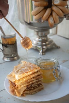 Drops of honey flow from a wooden spoon onto fresh pancakes, tea and a large samovar, the Russian tradition of celebrating Maslenitsa, a still life in the rays of the setting sun,
