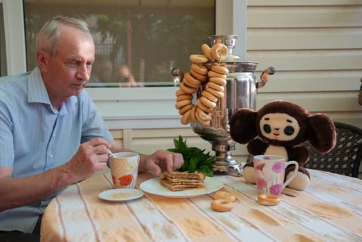 an elderly man enjoys a traditional Russian tea party with a samovar, pretzels, pancakes with honey in the company of a plush toy Cheburashka at the table,