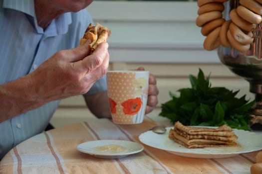 unrecognizable man dips pancakes in fresh acacia honey and holds them in his hand to take a bite, next to him is a samovar with buns and a stack of pancakes on a plate, celebrating Maslenitsa,