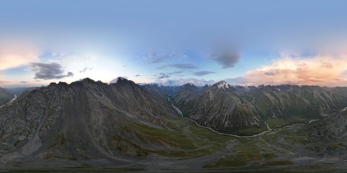 360 panorama of a mountainous area from a height. A drone view of sunrise in the mountains, orange clouds, snowy white mountain peaks, a green valley with river. Rocky terrain with cliffs. Khan Tengri