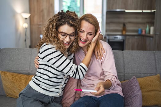 Two female friends are sitting on the couch in the living room, hugging and excited after getting their pregnancy test results.