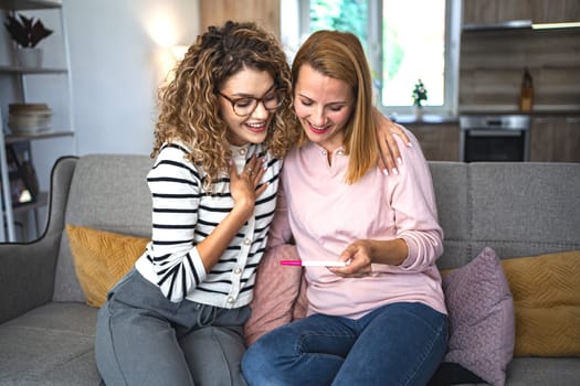 Two female friends are sitting on the couch in the living room, hugging and excited after getting their pregnancy test results.