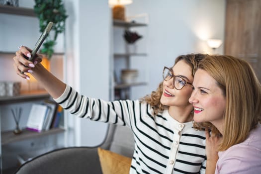 Two women are seated on a couch, holding a smartphone at arms length to take a selfie together. They are smiling while capturing the moment on the screen.