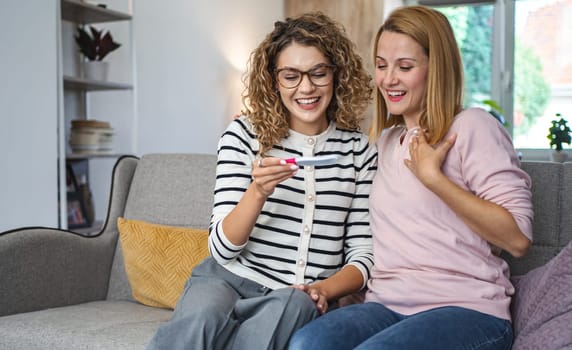 Two female friends are sitting on the couch in the living room, hugging and excited after getting their pregnancy test results.