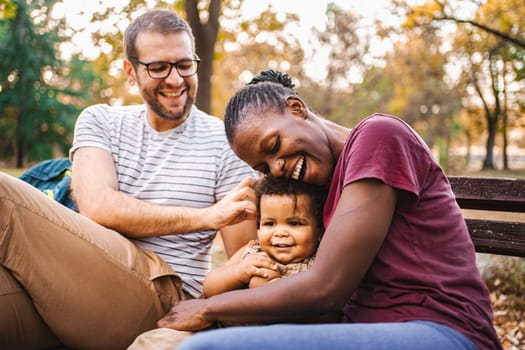 Mixed race couple is sitting on a park bench with their son.