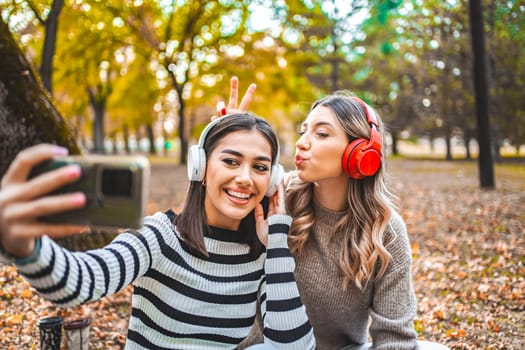 Two women are sitting on the ground, wearing headphones. They are focused on listening to music or audio together, possibly having a conversation or enjoying a podcast.