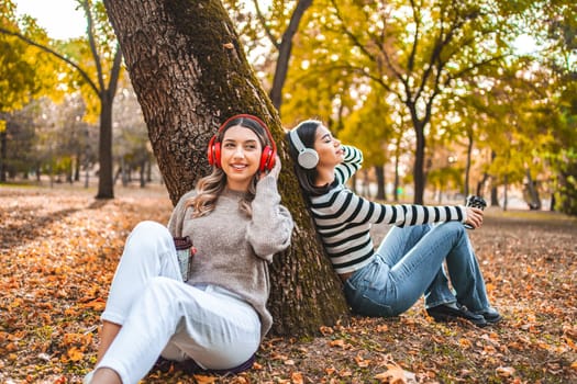 A woman is seated on the ground beside a tree, wearing headphones. She appears to be listening to music or an audio book, her attention focused on her device. The woman is dressed casually, and the setting seems to be outdoors in a park or garden.
