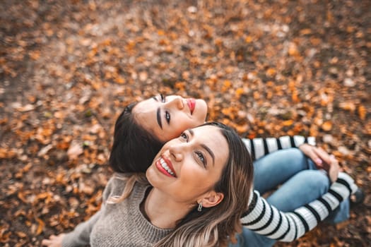 Two women are seen resting and reclining on top of a ground covered with fallen leaves. The women appear relaxed and comfortable as they enjoy the natural environment around them.