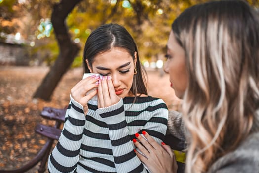 Two women are sitting on a park bench, one is comforting the other, while the other is sad and holds a handkerchief in her hand.