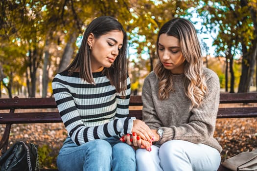 Two women are sitting on a park bench. They are facing each other, looking at each other, holding hands, comforting each other and giving each other comfort and support.