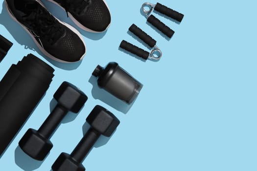 Black workout equipment, dumbbells, mat, hand grips, sneakers and water bottle on blue background. Fitness, sports items, healthy lifestyle concept. Place for your text