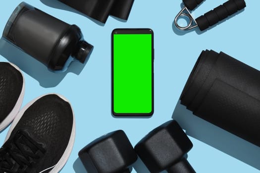 Smartphone mockup with green screen on the background of black fitness items, dumbbells, mat, sneakers, water bottle, sports elastic band. Demonstrating copy space for mobile application