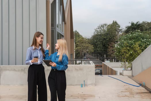 Two diverse women engaging in a lively conversation while holding a tablet and coffee outdoors in a contemporary setting.