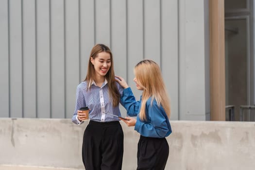 Two diverse women walking side by side and discussing ideas, with one holding a coffee cup and the other a tablet, in a modern outdoor setting.
