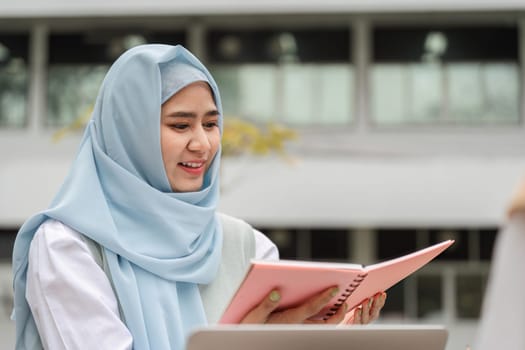 A young Muslim woman wearing a hijab studying and reviewing notes in her notebook outdoors at a university.