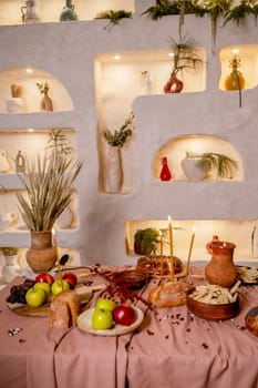 table set for Christmas or another holiday. candles, apples, bread, berries, dumplings are burning on the table. in the background is a decorative wall with lighting and clay pots. vertical photo