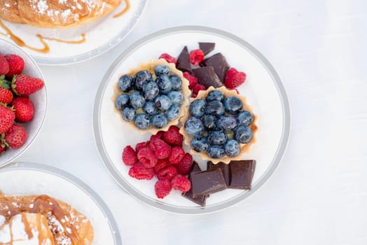 bright blueberries in tarlentines, raspberries, strawberries and chocolate next to them. fresh berries on a white tablecloth, also croissants with powdered sugar, chocolate pieces and strawberries