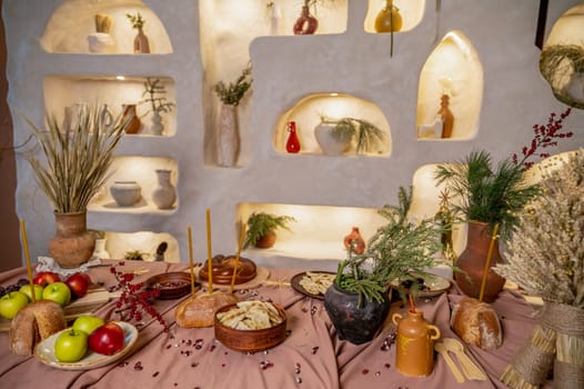 table set for Christmas or another holiday. candles, apples, bread, berries, grapes, dumplings are burning on the table. in the background is a decorative wall with lighting and clay pots. horizontal