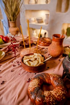 traditional table for a holiday. In the foreground there is a round loaf of bread, a loaf of bread, a plate with dumplings, a clay pot with ears of corn. In the background there is a decorative wall