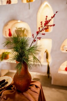 Still life with a clay pot of an old design with a pine branch and red berries. The vase is of a pleasant brown color. The photo is in warm shades, behind is a wall with backlighting.