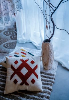Two decorative pillows lie on the floor next to a light decorative vase. There is also a striped rug on the floor. Light from a window covered with a curtain.