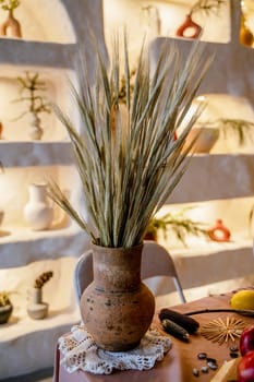 Still life with an antique clay pot and ears of wheat. The vase is of a pleasant brown color. The photo is in warm shades, behind is a backlit wall. There is a white handmade napkin on the table