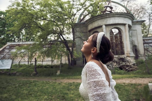 bride with smooth hairstyle and white headband in hair against old building background. brunette girl in white dress looking away. green trees, ruins of 18th century historic building in background