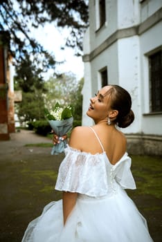 bride in a white dress with open shoulders and a bouquet of lilies of the valley. the brunette has a smooth hairdo gathered in a bun. behind is an old church of the 18th century. the girl is happy