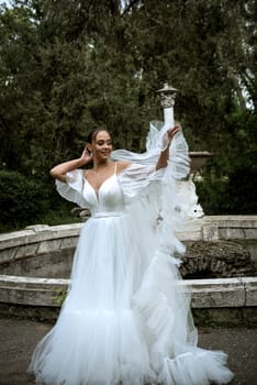 beautiful bride with long veil against the background of trees and old fountain. brunette girl in long white fluffy dress smiles and rejoices. bride throws long veil, behind old ruined fountain