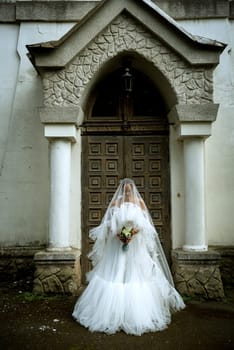 bride in a white dress covered with a chiffon veil and a bouquet of lilies of the valley. the girl stands straight upright in a long stylish dress against the backdrop of an 18th century church.