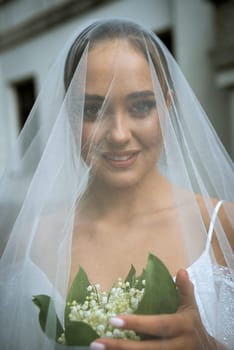 close-up of the bride in a white off-the-shoulder dress, with a veil, a bouquet of lilies of the valley. the brunette has a smooth hairdo tied in a bun. the bride is happy and smiling