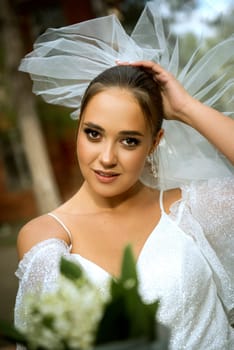 bride in a white dress with open shoulders, with a veil, a bouquet of lilies of the valley. the brunette has a smooth hairstyle gathered in a bun. she is looking at the camera,