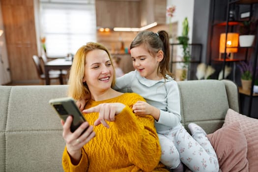 Mother and daughter sitting on couch in living room, smiling and spending time looking at mobile phone