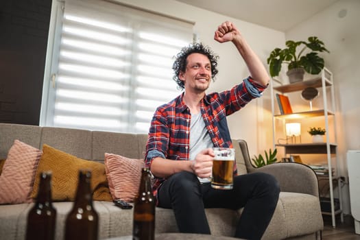 Mid adult man is happily cheering while watching sports on television and drinking beer at home