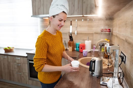 A woman is standing in the kitchen with a towel on her head. She makes herself a coffee.