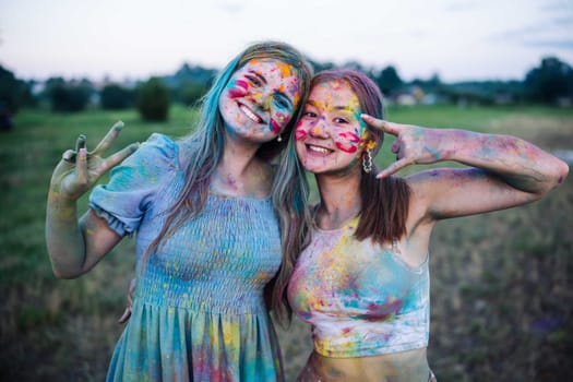 Two young women, covered in vibrant holi powder, gesture victory signs, celebrating friendship and the joy of the festival in a field