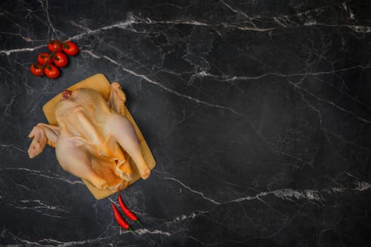 The chicken carcass, butchered and plucked, lies on a wooden plank against a background of black marble, surrounded by cherry tomatoes and hot red peppers. The concept of cooking, poultry meat, dietary dishes. High quality photo