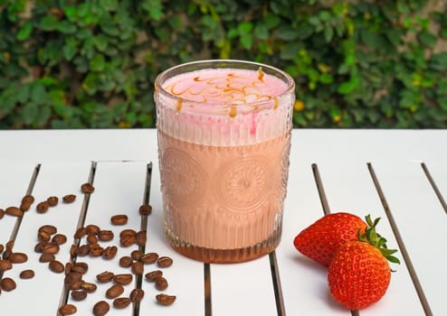 Strawberry latte with fresh strawberry and coffee beans on white wooden table against green leaves with copy space