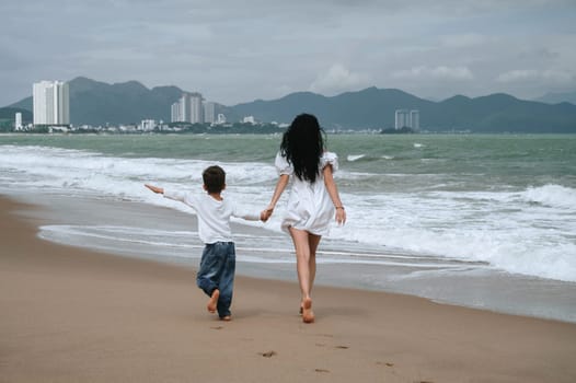 back of happy mom and son walking along the beach by the sea on summer vacation