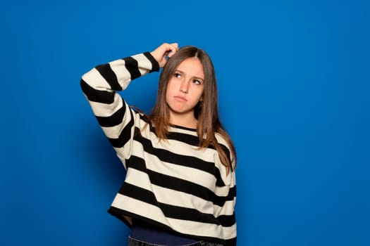 Hispanic teenage girl about 15 years old wearing a striped sweater and a puzzled facial expression, scratches her head in frustration, isolated over a blue background. Hmm, I don't remember.