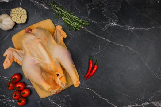 The butchered raw chicken lies on a wooden cutting board against a background of black marble, surrounded by tomatoes, red hot peppers, rosemary and garlic. The concept of cooking, diet menu, ingredients. High quality photo
