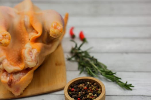 A close-up side view of a butchered raw chicken lying on a wooden cutting board, on the side there is rosemary, hot red pepper, and a wooden bowl made of a mixture of peppers against a white wood background with space for text. The concept of cooking, diet menu, ingredients. High quality photo