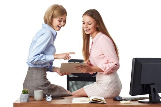 Two office female workers discuss project, sitting on desk with clipboard in office