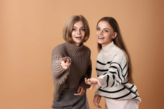 Two women friends together in casual clothes point index finger aside on beige background. Surprised blonde woman and her friend