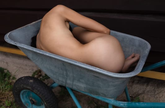 Young nude woman posing lying in a construction wheelbarrow at a country house construction site