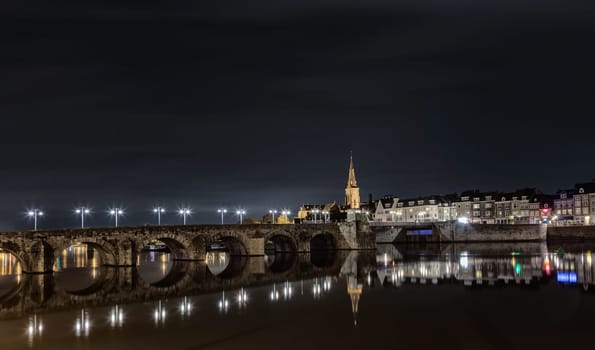 Riverside night scene in Maastricht, Netherlands with old bridge over river Maas and illuminated Saint Martin church.