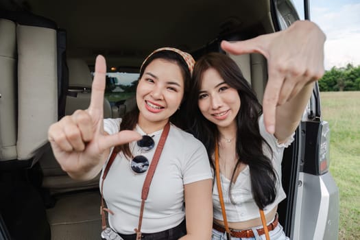 Two young Asian women in white shirts and jeans sit in the back of their new car, lounging on the field. holidays and traveling.